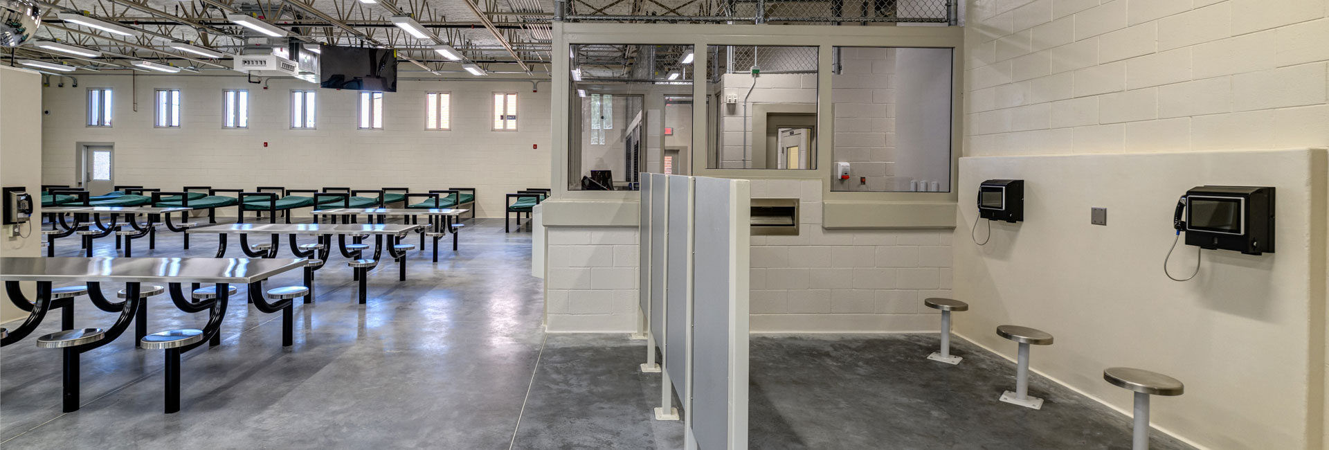 The side view of the room at Volusia County Branch Jail with steel bench and stools, back metal cots, a wall mounted phone section and the glass box of the officer station, built by the Ajax Building Company.