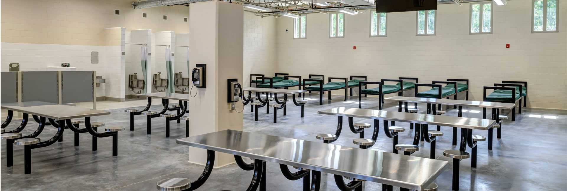 Dorm of the Volusia County Branch Jail with cots along the wall, metal benches and stools in center and pay phones on pillars in center of room, built by Ajax Building Company.