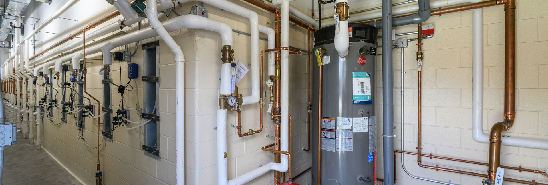 View of the back wall of the plumbing with a water heater or boiler unit with white PVC and bronze pipes at the Volusia County Branch Jail built by the Ajax Building Company.